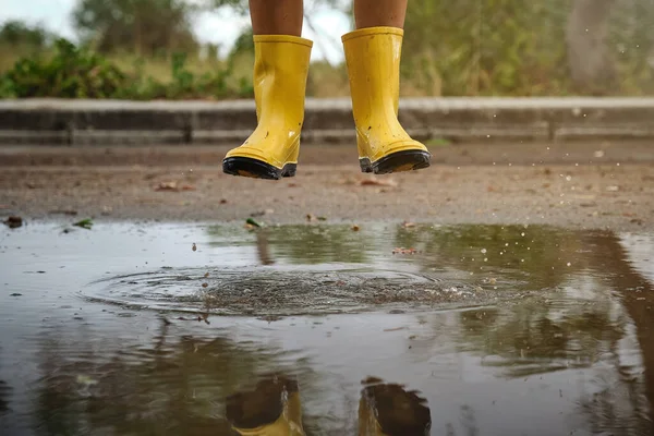 Crop anonymous child in yellow rubber boots jumping in puddle on muddy road against green trees on cloudy day