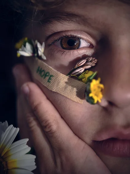 Half of face of tranquil boy with flowers on face and plaster with Hope inscription looking at camera while touching cheek