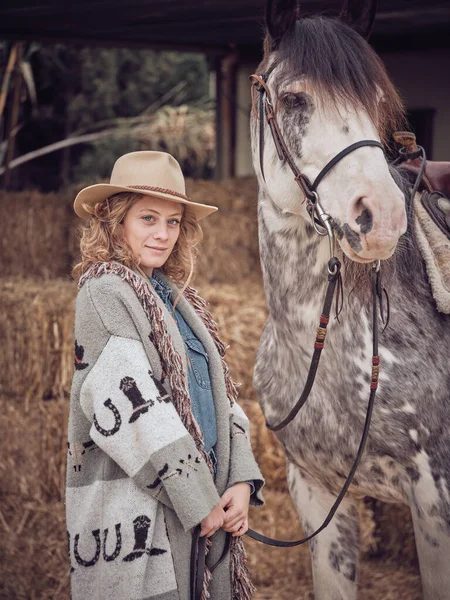Charming woman in cowgirl outfit looking at camera and purebred stallion in bridle standing against haystacks