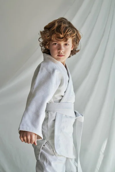 Duygusuz Kıvırcık Saçlı Beyaz Kimonolu Çocuk Perdenin Yanında Durmuş Yumruğunu — Stok fotoğraf