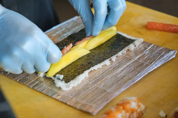 レストランのキッチンで働く中で 海苔にマンゴーとサーモンを使った新鮮な寿司を作る手袋で顔のない料理人 — ストック写真