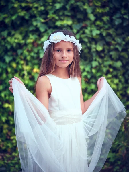 庭に立ってカメラを見ている白いドレスと花輪を身に着けているかわいい少女の笑顔の肖像 — ストック写真