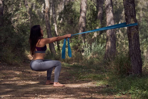 Full body side view of long haired female athlete squatting with resistance band during fitness training in sunny summer park
