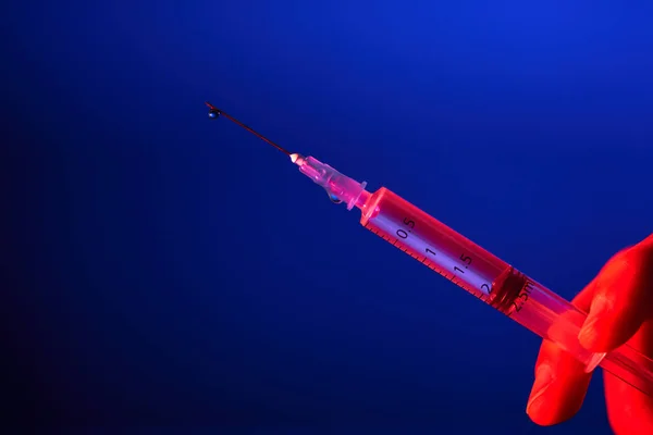 Medical syringe with needle full of vaccine for COVID19 in hand of anonymous doctor against blue background in studio with red light
