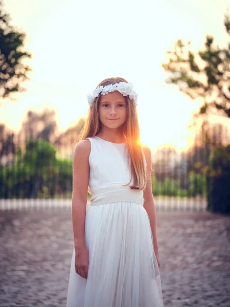 幸せなPreteen子供で白いドレスと花の花輪笑顔とカメラを見ている間に日没の空と緑の木に対して立って — ストック写真