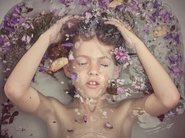 明るい浴室のカラフルな小さな花と透明な浴槽の下に横たわっている閉じた目を持つ落ち着いた男の子のトップビュー — ストック写真