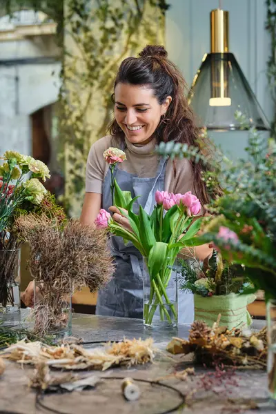 Professional female florist in apron standing at table with various plants and flowers and looking at blossoming carnation while working in floral shop