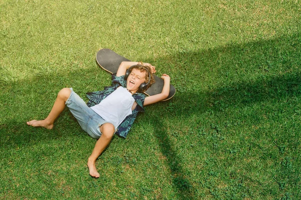 草の芝生の上にスケートボードに横たわっている間 興奮した前の子供の叫びとヘッドフォンで音楽を聴くの上から — ストック写真