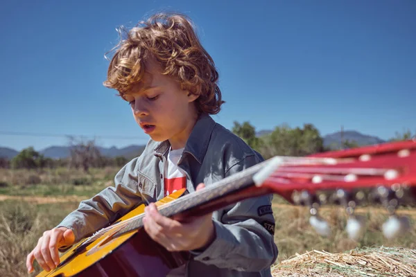 田舎の雲のない青空の下で芝生のフィールドに座ってアコースティックギターを演奏カジュアルな服でかわいいブロンドの髪を持つ集中した小さな少年 — ストック写真
