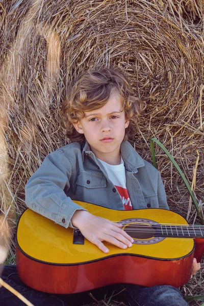 田舎の干し草のロールの近くにアコースティックギターと座っているかわいい髪の男の子と離れて見る — ストック写真