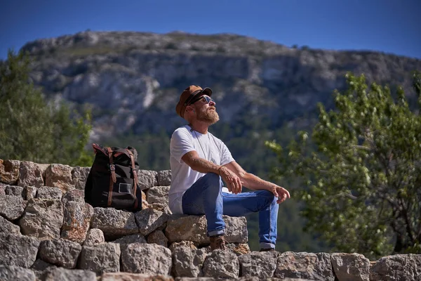 サングラスや帽子で髭を生やした男性観光客の完全な体は リュックサックの近くに石の階段に座って 晴れた日の間に絵のように山岳風景を楽しむ — ストック写真