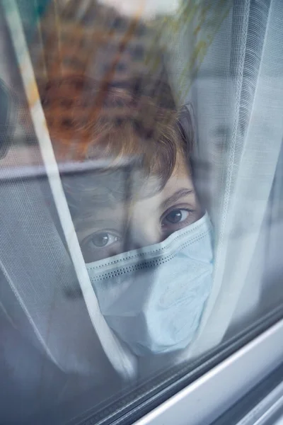 コロナウイルスのパンデミック中にカーテンで窓の後ろに立っている間 カメラを見ている保護医療マスクを着用している19歳の子供のガラスビューを通して — ストック写真