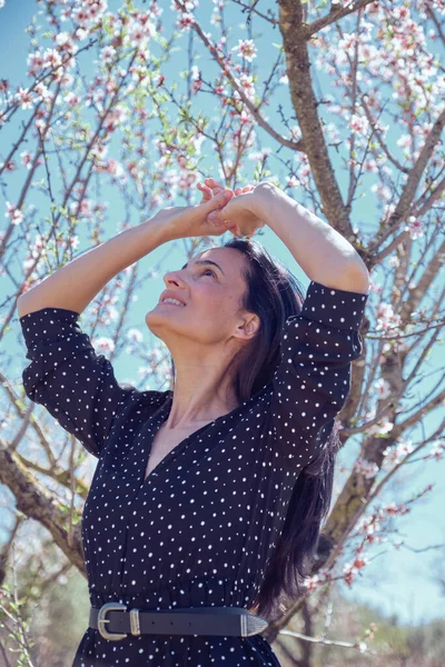 ドットドレスで笑顔の女性の下から ベルトを見上げて緑の木の下に立って 葉で覆われ 雲のない青空の下でピンクの花を咲かせます — ストック写真