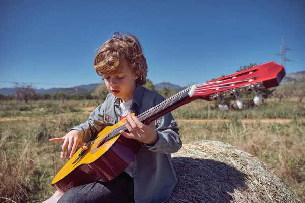 かわいいブロンドの髪のカジュアルな服を着て 田舎の雲のない青空に対してアコースティックギターを演奏する男の子 — ストック写真