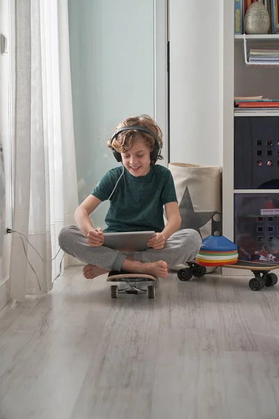 Celé Tělo Usměvavý Chlapec Sluchátkách Sedí Skateboardu Zkříženýma Nohama Sleduje — Stock fotografie