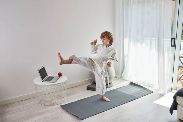 Full body of focused child in kimono with white belt standing on sports mat and doing judo while watching tutorial on laptop in living room at home