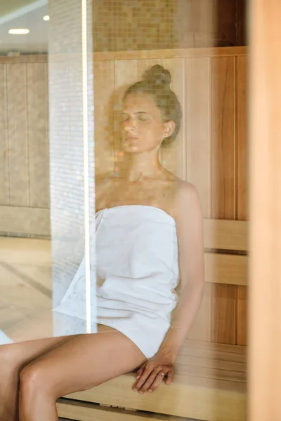 レクリエーションセンターで週末を過ごす間 目を閉じてサウナで木製のベンチに休んで白いタオルで若い女性のガラスを通して — ストック写真