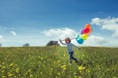 Küçük tatlı çocuk mavi gökyüzüne karşı renkli balonlarla oynarken çayırlarda koşuyor.