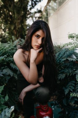 Muhteşem bahçedeki yeşil çalıların yanında durmuş kameraya bakan güzel genç bir kadın.