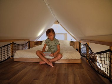 Yatak odasında bacak bacak bacak üstüne atmış, çıplak ayaklı bir çocuk. Elinde defterle rahat bir yatağın üstünde glamping çadırının altında oturuyor.
