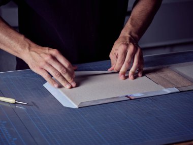 Stüdyodaki çalışma masasında yeni bir kitap yaparken, isimsiz bir erkek kitap ciltçisi karton kitap kapağını kağıtla yapıştırıyor.