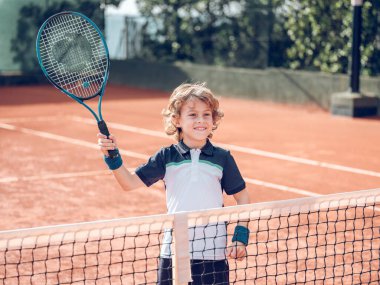 Sarışın, kıvırcık saçlı, mutlu bir çocuk raketle ağların yanında durup kil sahada tenis maçında topa vurmaya hazırlanırken gülümseyip bakışlarını kaçırıyor.