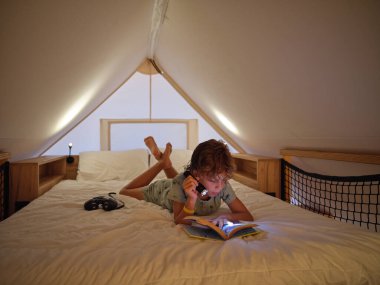 Gündelik giyinmiş çıplak ayaklı bir çocuk. Parlak el feneri takıyor ve rahat beyaz yatakta yatarken ilginç kitaplar okuyor.