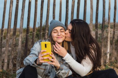 Uzun siyah saçlı, günlük kıyafetleriyle kırsal alanda ahşap bir yolda oturan ve akıllı telefon kullanarak en iyi arkadaşının yanağına sarılıp öpen genç bayan.