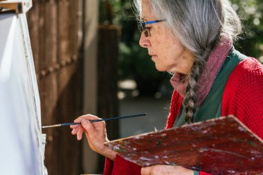 Yazın güneşli bir günde arka bahçede tuvale resim yapan yaşlı kadın ressamların yan görüntüsü