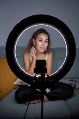 Akıllı telefondan video kaydederken yüzünü gösteren kıvırcık saçlı bir kadın blogcu. Üç ayaklı ve ışıklı bir lamba kullanıyor.
