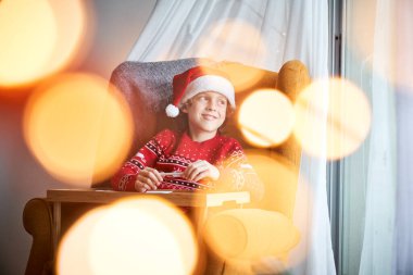 Noel Baba şapkalı sevimli çocuk çizim sırasında elinde kalem tutarken koltuğunda oturuyor ve başka tarafa bakıyor.