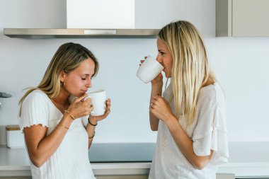 Bir fincan kahveden içen olumlu bayan arkadaşlar mutfakta modern mutfak malzemeleriyle birbirlerine bakıyorlar.