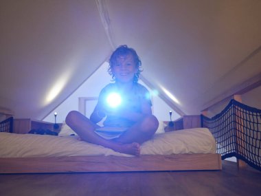 Gündelik defterleri olan neşeli bir çocuk. Parlayan el feneri takıyor ve Mansard 'da yatakta otururken kameraya bakıyor.