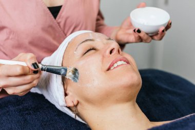 Modern güzellik salonundaki cilt bakımı sırasında kadın müşterinin yüzüne nemlendirici yüz maskesi uygulayan tanınmamış kozmetik uzmanı.