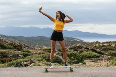 Asfalt yolda uzun tahta üzerinde dururken akıllı telefondan selfie çeken etnik bir kadın.