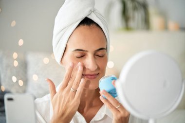 Kafasında havlu sarığı olan mutlu kadın blogcu vlog için akıllı telefondan video çekerken gözleri kapalı nemlendirici yüz kremi uyguluyor.