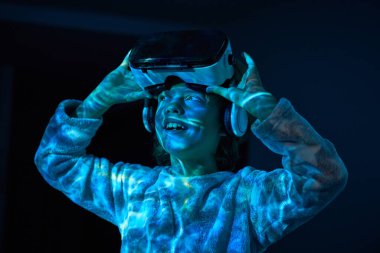 Sıcak pijama giymiş, mutlu bir şekilde gülümseyen ve karanlık odada VR kulaklığını çıkarırken gözlerini kaçıran küçük hayret verici bir çocuk.