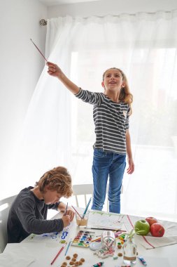 Sandalyede elinde fırçayla gülümseyen bir kız ve üzerinde çeşitli tatlılar bulunan akvaryum boyalarıyla kağıt bir çarşaf çizen bir çocuk.