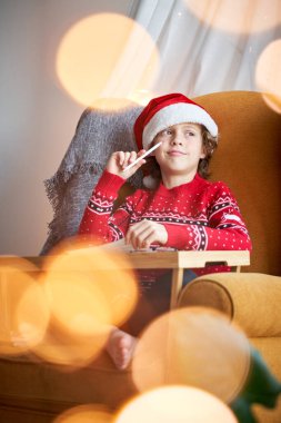 Kalemle çizen, koltukta oturup Noel Baba şapkası takan düşünceli, sevimli bir çocuk.