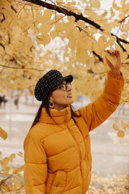 Şık, sıcak giysiler içinde güzel genç bir bayan. Gülümseyen ve sarı yapraklara dokunan şehir caddesinde muhteşem bir sonbahar günü.