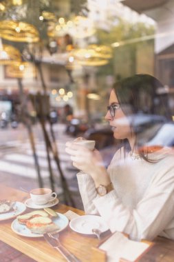 Moda kıyafetleri içinde güzel bir bayan elinde bir fincan sıcak içecekle ve rahat bir kafede pencerenin arkasında otururken başka tarafa bakıyor.