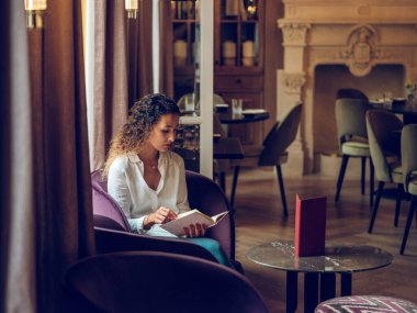 Odaklanmış kıvırcık saçlı genç bir kadın kanepede oturuyor ve ilgiyle kitap okuyor.