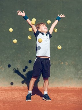 Uzun boylu, sarı saçlı, aktif giysili, mutlu bir şekilde gülümseyen, gözleri kapalı, tenis antrenmanından sonra kollarını kaldıran sevimli bir çocuk.