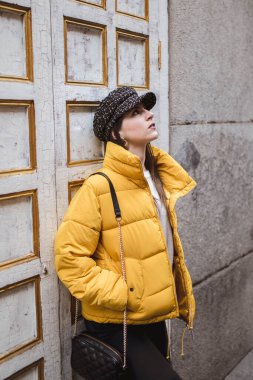 Şehir caddesinde yıpranmış duvarın yanında dikilirken şık kıyafetli güzel bir kadın başka tarafa bakıyor.