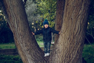 Doğada ağaç gövdesinde dikilirken şapkalı ve dış giysili bir çocuğun tüm vücudu merakla yüzünü çeviriyor.