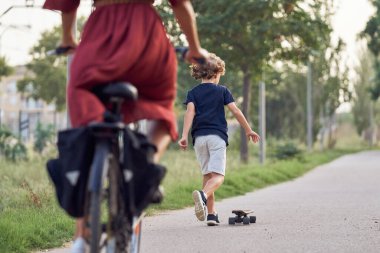 Tanımlanamayan bir çocuğun arka görüntüsü günlük giysiler içinde kaykay sürerken asfalt yolda parkta bisikletli bir kadının arkasında.