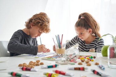 Karmaşık masadaki boya fırçalarıyla, renkli boyalarla ve aydınlık odada dağınık sanat malzemeleriyle resim yapan ciddi kardeşler.