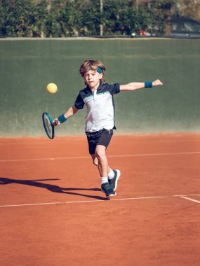 Spor giyim ve saç bandı giymiş, güneşli bir günde kortta raketle tenis oynayan küçük bir çocuğun tüm vücudu.