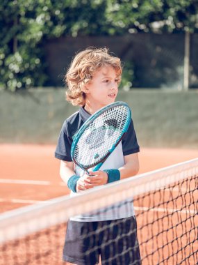 Spor kıyafetleri ve bileklikleriyle kıvırcık sarı saçları olan, tenis kortunun yanında raket tutan ve dışarıda antrenman yaparken gözlerini kaçıran sevimli bir çocuk.