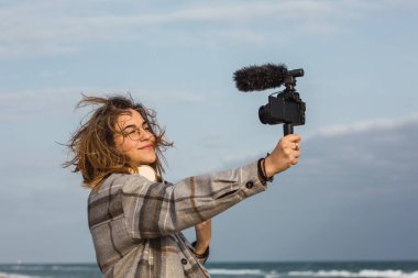 Sıcak kareli gömlekli neşeli genç bayan blogcu kumlu sahilde gülümserken bulutlu gökyüzüne karşı kamerada vlog kaydediyor.